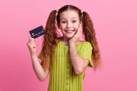 Photo portrait of cute little girl hold credit card amazed dressed stylish green clothes isolated on pink color background.