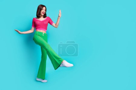 Full size photo of positive girl wear pink knit top green trousers dancing look empty space show shoe isolated on blue color background.