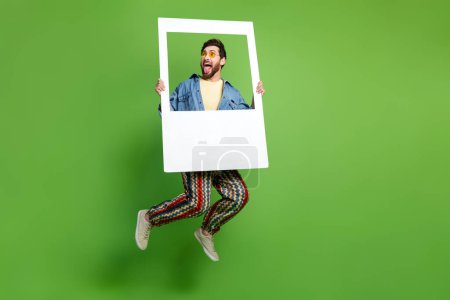 In voller Länge Foto von beeindruckt aufgeregt Kerl gekleidet Jeans-Shirt springen hoch halten Fotorahmen isoliert grüne Farbe Hintergrund.