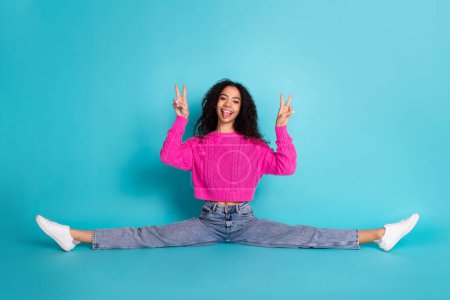 Full size photo of pretty teen girl split sit floor v-sign wear trendy pink outfit isolated on aquamarine color background.