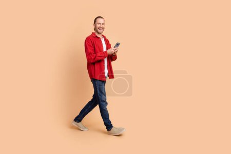 Full Size Profilfoto von netten jungen Mann halten Telefon zu Fuß leeren Raum tragen rotes Hemd isoliert auf beige Farbhintergrund.
