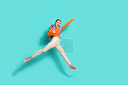 Photo pleine grandeur de fille extatique habillée sac à dos chemise orange sur l'épaule livre volant levant poing isolé sur fond de couleur sarcelle.