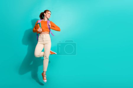 pleine taille photo de fille diligente habillée orange chemise sac à dos tenir livre volant look vide espace isolé sur fond de couleur sarcelle.