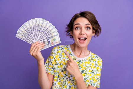 Porträt der hübschen jungen Frau zeigen Finger Dollarscheine tragen T-Shirt isoliert auf lila Hintergrund.