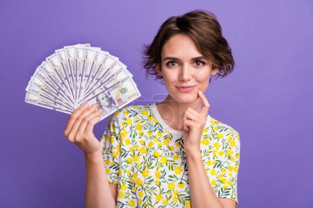 Porträt der hübschen jungen Frau halten Bargeld denken tragen T-Shirt isoliert auf lila Hintergrund.