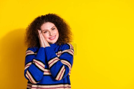 Porträt von niedlichen schönen Mädchen mit welliger Frisur tragen Strickpullover mit Handflächen auf Wangenknochen isoliert auf gelbem Hintergrund.