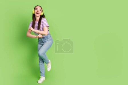 Foto de longitud completa de hermosa joven mirada espacio vacío audífonos de baile vestido de rayas violeta prenda aislada sobre fondo de color verde.