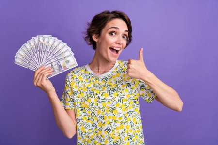Retrato de una mujer joven y bonita con dinero mostrar el pulgar hacia arriba usar camiseta aislada sobre fondo de color púrpura.