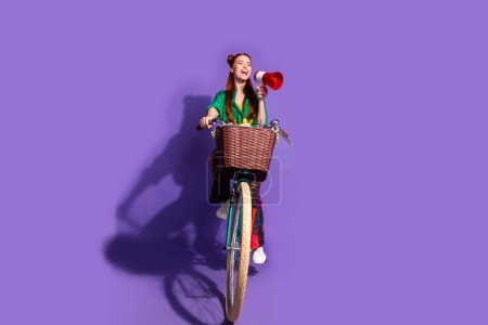 Full length photo of adorable girl wear shirt driving bicycle scream in megaphone look empty space isolated on violet color background.