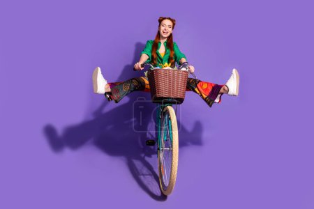Full size photo of crazy carefree woman dressed green shirt vintage pants hold legs up riding bike isolated on purple color background.