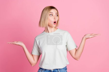Portrait of impressed girl with bob hair wear white t-shirt arms comparing products look empty space isolated on pink color background.