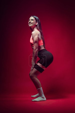 Young sportive attractive caucasian athlete woman posing in pink tank top and black sauna shorts fitness outfit. Studio shooting with pretty slim fit brunette doing aerobic stretching with rubberbands