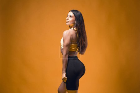 Young sportive attractive caucasian athlete woman posing in yellow tank top and black sauna shorts fitness outfit. Studio shooting of pretty slim fit brunette doing aerobic stretching with rubberbands