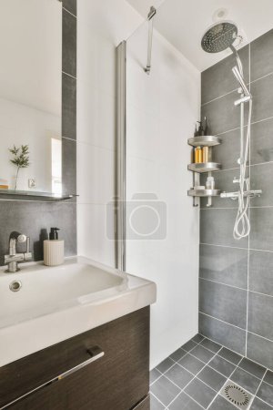 Foto de Un moderno cuarto de baño con azulejos grises y accesorios blancos en el cabezal de la ducha está montado en la pared por encima del fregadero - Imagen libre de derechos