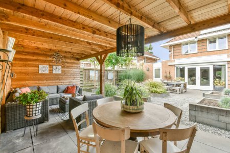 Foto de Un patio con sillas, mesas y plantas en la mesa frente a una sala de estar al aire libre que tiene paredes con paneles de madera - Imagen libre de derechos