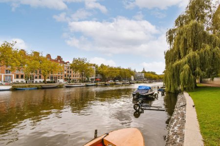 Téléchargez les photos : Amsterdam, Pays-Bas - 10 Avril, 2021 : quelques bateaux sur l'eau et des bâtiments en arrière-plan, avec des arbres bordant les rives de la rivière à droite - en image libre de droit