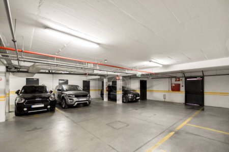 Photo for Two cars parked in a parking space with yellow lines painted on the floor and white walls, along with black doors - Royalty Free Image