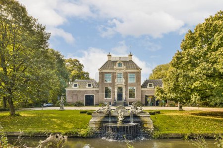 Photo for A large house with a fountain in the front and trees surrounding it on a sunny, blue cloudy sky day - Royalty Free Image