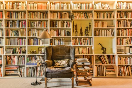 Foto de Ámsterdam, Países Bajos - 10 de abril de 2021: una silla delante de un estante de libros con muchos libros y una lámpara sentada junto a la silla - Imagen libre de derechos