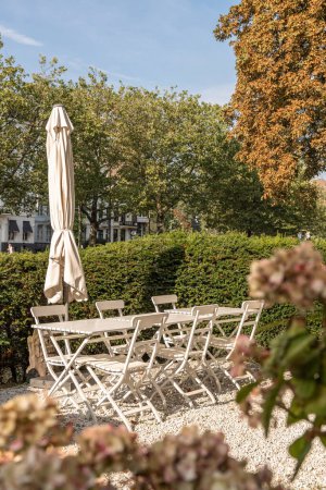 Téléchargez les photos : Une table et des chaises dans un jardin avec un parasol sur la table, entourée de buissons et d'arbres par une journée ensoleillée - en image libre de droit