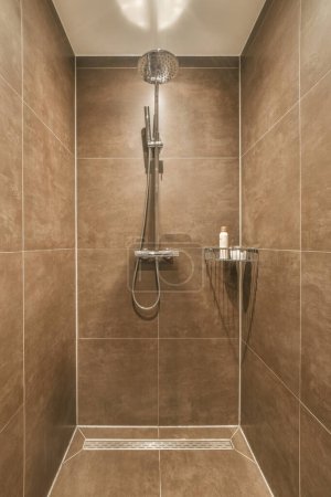 Photo for A shower room with brown tiles on the walls and tile around the tub, hand held by an adjustable shower head - Royalty Free Image
