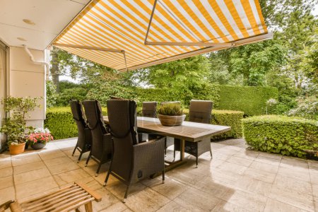 Téléchargez les photos : Un espace repas extérieur avec table et chaises sous un grand auvent à rayures jaunes sur le patio, entouré d'arbres verts luxuriants - en image libre de droit