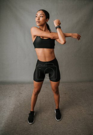 Foto de Mujer Multi étnica de pie y muy centrada haciendo sus ejercicios de estiramiento del brazo con su reloj de bits en forma mientras está en el estudio. Foto de alta calidad - Imagen libre de derechos