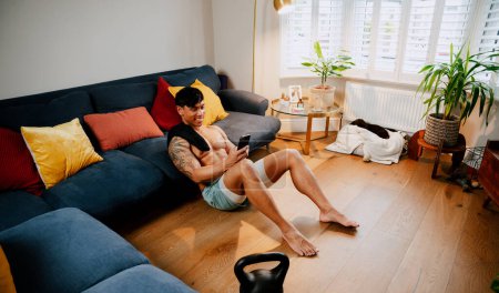 Téléchargez les photos : Tatoué asiatique mâle sur son téléphone portable assis sur son plancher en bois - en image libre de droit