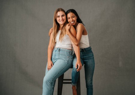 Téléchargez les photos : Deux amies souriant joyeusement partageant haut un tabouret là-dedans jeans et t-shirts. Photo de haute qualité - en image libre de droit