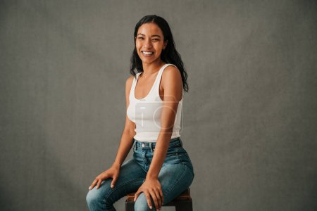 Photo for Multiethnic female smiling and sitting on a stool hand on both knees in the studio . High quality photo - Royalty Free Image