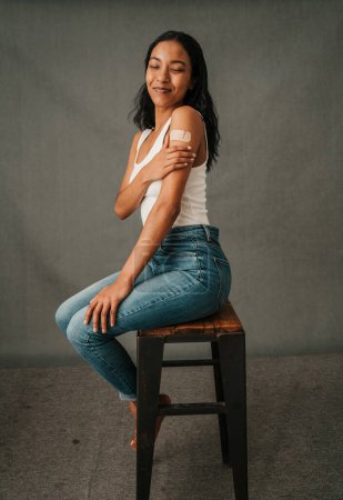 Foto de Joven hembra multiétnica mirando la tirita en su brazo mientras está sentada en una silla alta. Foto de alta calidad - Imagen libre de derechos