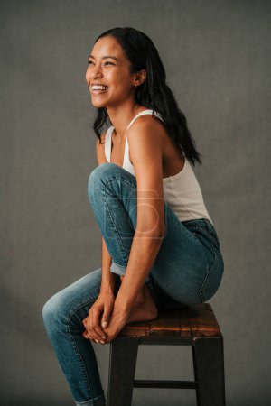 Téléchargez les photos : Femme multiethnique avec les cheveux noirs assis et riant dans le studio pieds nus dans son jean. Photo de haute qualité - en image libre de droit