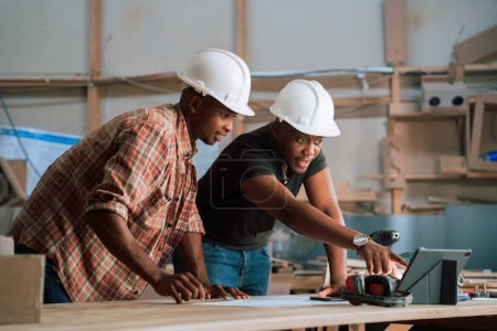 Téléchargez les photos : Deux artisans africains avec là-bas des chapeaux durs sur le travail sur leurs plans et là tablette dans leur usine. Photo de haute qualité - en image libre de droit