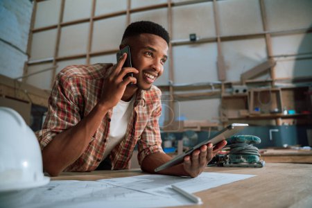 Photo for Smiling young African male uses cellphone while holding tablet in woodwork warehouse . High quality photo - Royalty Free Image