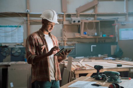 Photo for Young African male with hardhat stands in woodwork factory while using tablet . High quality photo - Royalty Free Image