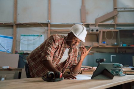 Téléchargez les photos : Jeune homme africain regarde les dessins tout en utilisant la tablette et le téléphone dans l'usine de menuiserie. Photo de haute qualité - en image libre de droit