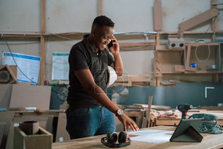 Téléchargez les photos : Homme africain utilise tablette pour aider la conception dans une usine de menuiserie tout en parlant au téléphone. Photo de haute qualité - en image libre de droit