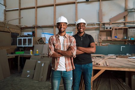 Téléchargez les photos : Des collègues africains masculins portant des casques durs sourient dans une usine de menuiserie les bras croisés. Photo de haute qualité - en image libre de droit