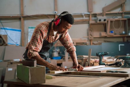 Téléchargez les photos : Un homme africain concentré portant des cache-oreilles mesure un morceau de bois pour un projet de menuiserie. Photo de haute qualité - en image libre de droit