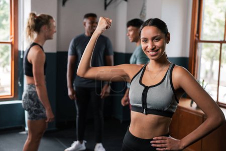 Photo for A woman in an undershirt and brassiere is smiling while flexing her arm and thigh muscles in front of a window at the gym - Royalty Free Image