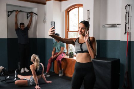 Photo for A woman is capturing a selfie in the gym using her phone, showcasing her toned muscles and athletic physique. She is wearing shorts and her hand is extended, flexing her arm and leg muscles - Royalty Free Image