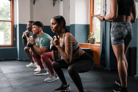 Téléchargez les photos : Un groupe d'individus travaillent dans la salle de gym, accroupi avec des haltères pour renforcer leurs muscles des jambes et des articulations pendant leur temps libre - en image libre de droit