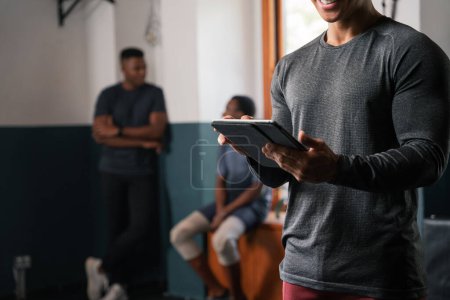 Photo for A man in formal wear is holding a tablet in a gym building. He is listening to music using audio equipment in the room while working out his thighs - Royalty Free Image