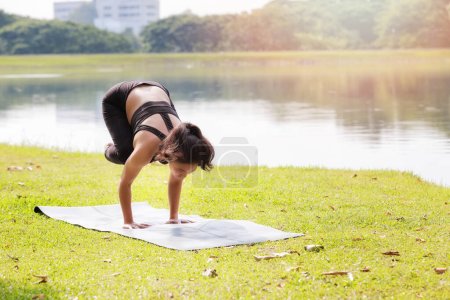 Asian woman practicing yoga doing Crane (Crow) Pose, Bakasana pose on the mat beside a lake in outdoor park.