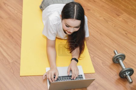 Hermosa joven mujer viendo videos tutoriales de fitness en línea en el ordenador portátil mientras se acuesta en el piso en la sala de estar.