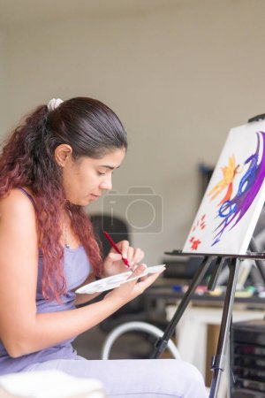 Téléchargez les photos : Femme adulte latine peignant à la maison le week-end. artiste passe-temps faire des œuvres d'art acrylique sur toile. détente et tranquillité - en image libre de droit