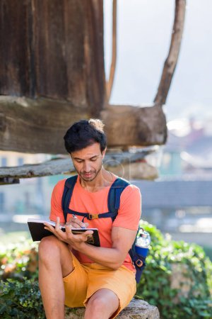 Foto de Hombre mochilero adulto medio asiático del sur sentado con cuaderno diario en Cadavedo Asturias. Estilo de vida al aire libre retrato adulto haciendo turismo con portátil - Imagen libre de derechos