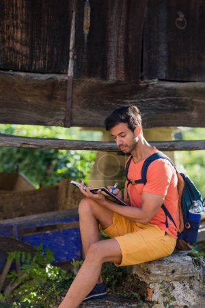 Foto de Hombre mochilero adulto medio asiático del sur sentado con cuaderno diario en Cadavedo Asturias. Estilo de vida al aire libre retrato adulto haciendo turismo con portátil - Imagen libre de derechos