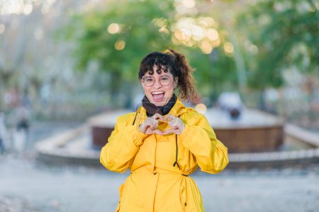 Photo for Venezuelan happy and friendly middle-aged woman on yellow winter raincoat outdoors. IWD2024 feminist make hand symbol - Royalty Free Image