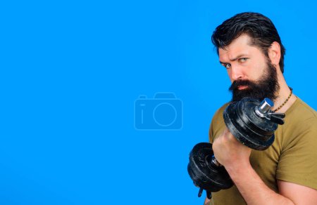Photo for Serious bearded man with dumbbell in hands. Athletic muscular sportsman lifting dumbbell in sport club. Strong handsome bodybuilder at workout in fitness gym. Weight training. Copy space. Advertising - Royalty Free Image